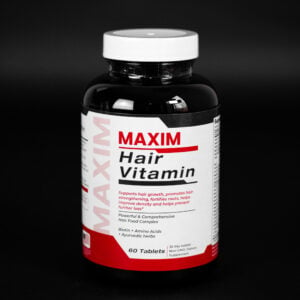 A product shot of a black plastic bottle of Maxim Hair Vitamin supplements against a black background. The bottle has a white label with red and black text. The text reads "MAXIM" vertically on the left side of the label, and "MAXIM Hair Vitamin" in red and black in the center. Below that, the label states "Supports hair growth, promotes hair strengthening, fortifies roots, helps improve density and helps prevent further loss*". It also mentions "Powerful & Comprehensive Hair Food Complex", "Biotin + Amino Acids + Ayurvedic herbs", "60 Tablets", and "30 day supply Non-GMO, Dietary Supplement". The bottle has a white cap.