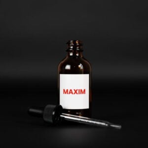 A close-up studio shot shows a brown glass bottle with a white label that reads "MAXIM" in red capital letters. The bottle is positioned in the center of the frame against a solid black background. In front of the bottle, a glass dropper with a black rubber bulb is lying horizontally. The lighting is soft, creating subtle reflections on the glass surfaces.